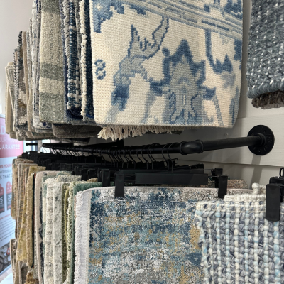 Double-level black metal racks displaying hanging rug samples in shades of blue, gray, and beige, with various patterns and textures, against a white background.